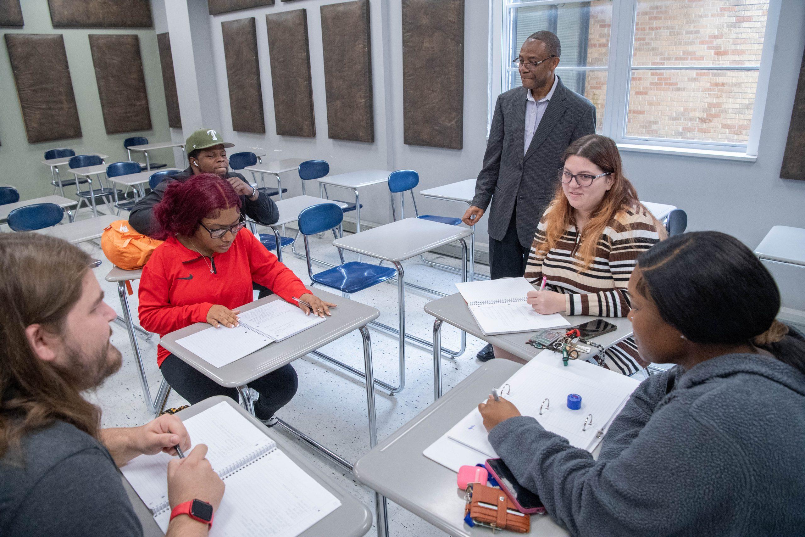 Five students with open notebooks sitting in a group in class, with a professor standing near them.