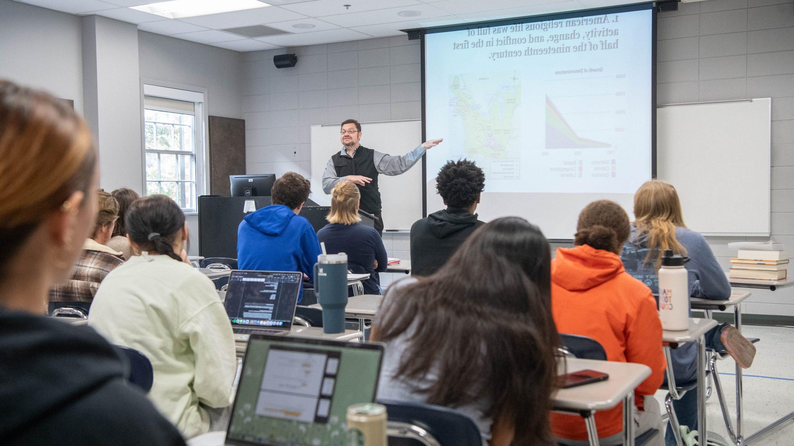 Professor giving lecture, with a PowerPoint next to him, in front of students in a classroom.