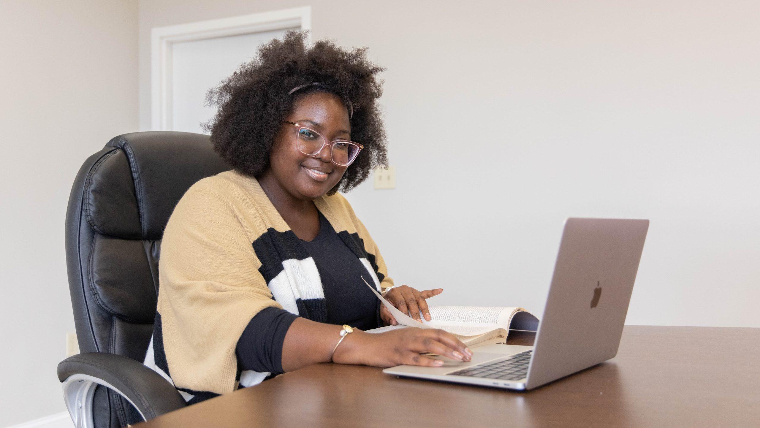 A woman sitting at a table in a school conference room, smiling with a computer in front of her.