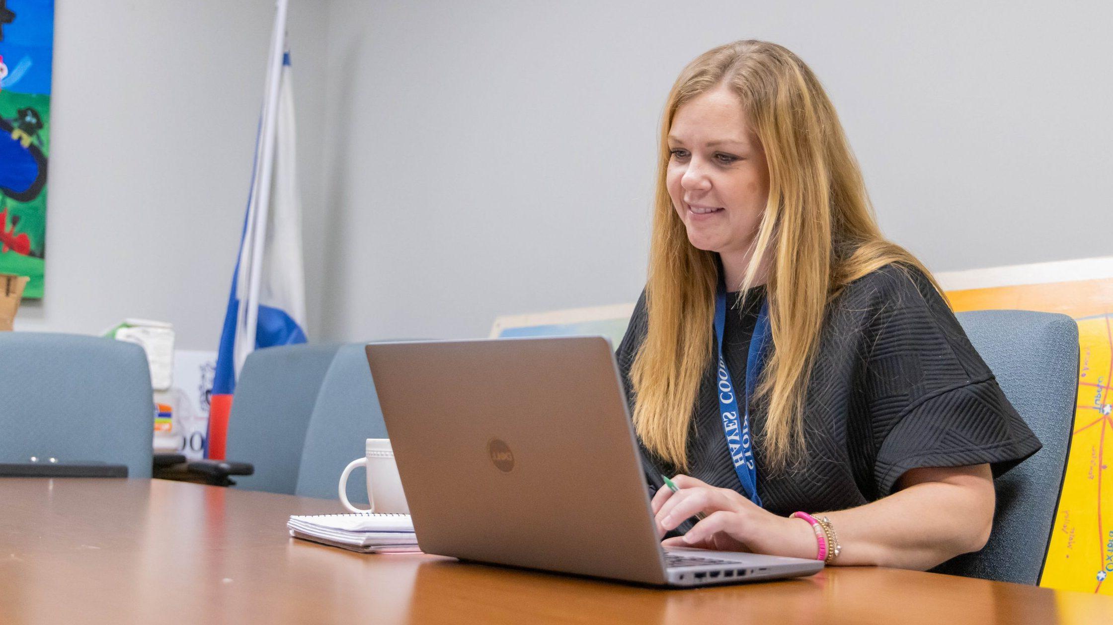 A woman sitting at a table in a conference room, smiling while looking at her computer.