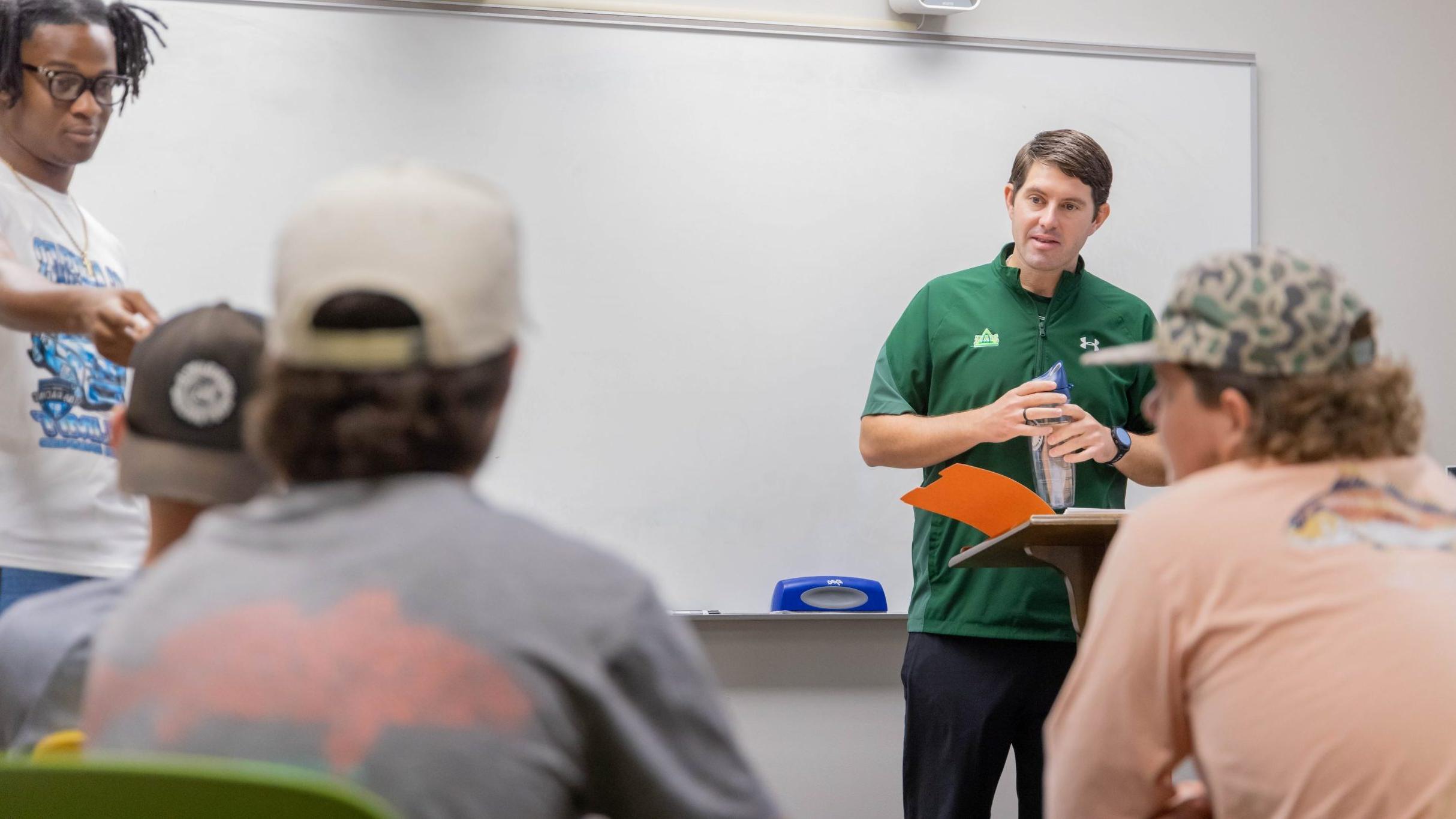 An instructor and a student standing in front of a classroom, with three students sitting at desks, facing forward towards them.