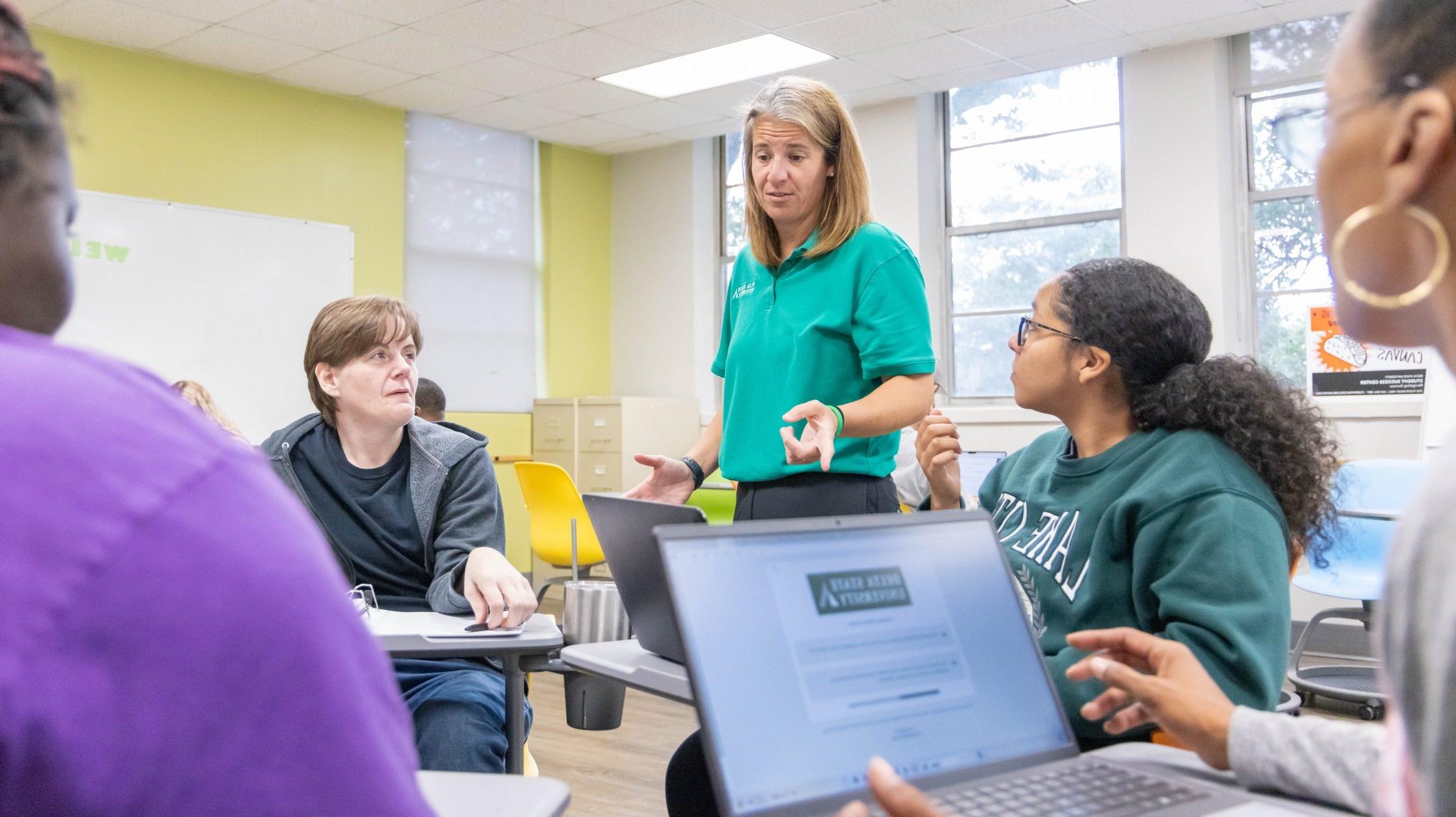 Instructor helping a group of students sitting at desks organized in a circle. Each student is looking at the instructor.